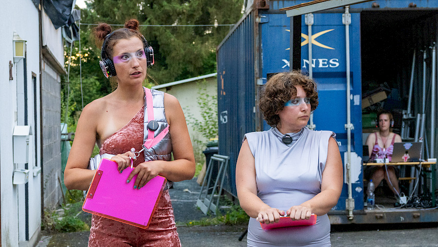Szenenfoto aus SUPER BADASS WOMEN, eine Schauspielerin hat Kopfhörer auf andere hält Klemmbrett