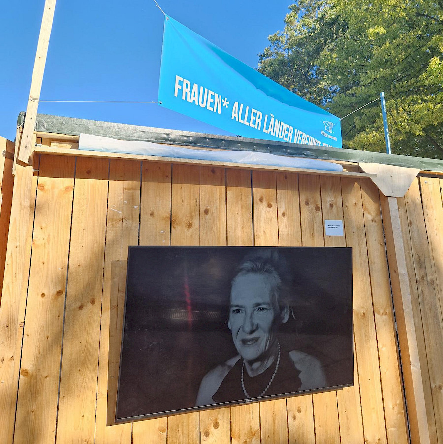 Alter, kleiner Container von Außen. Angebrachter Fernseher zeigt Videoinstalation, auf dem Dach das Banner darauf steht " Frauen* aller Lander vereinigt euch"