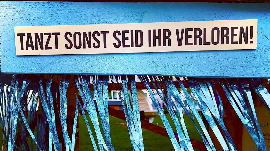 Ein blau bemaltes Holzbrett, mit Lametta, darauf steht "Tanzt sonst seid ihr verloren"